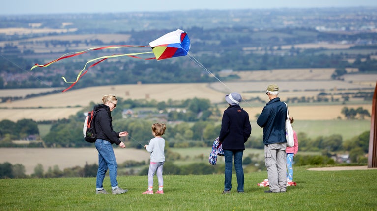 Large family of adults and children standing on top of hill watching while elderly man flies a kite